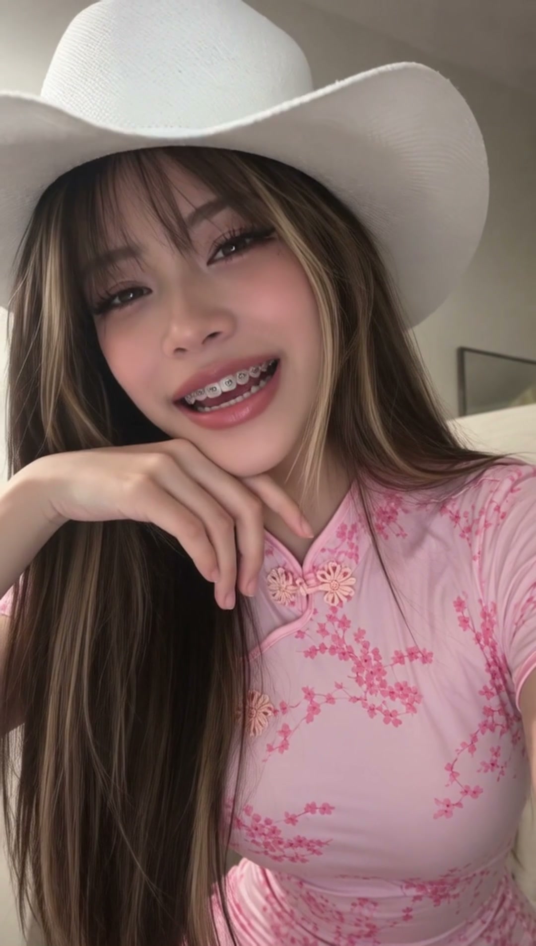 AI generated girl Primela Rose wearing a white cowboy hat, pastel pink cherry-blossom Chinese qipao dress, blonde face-framing highlights, and metallic braces singing a Japanese J-Pop song in close-up POV