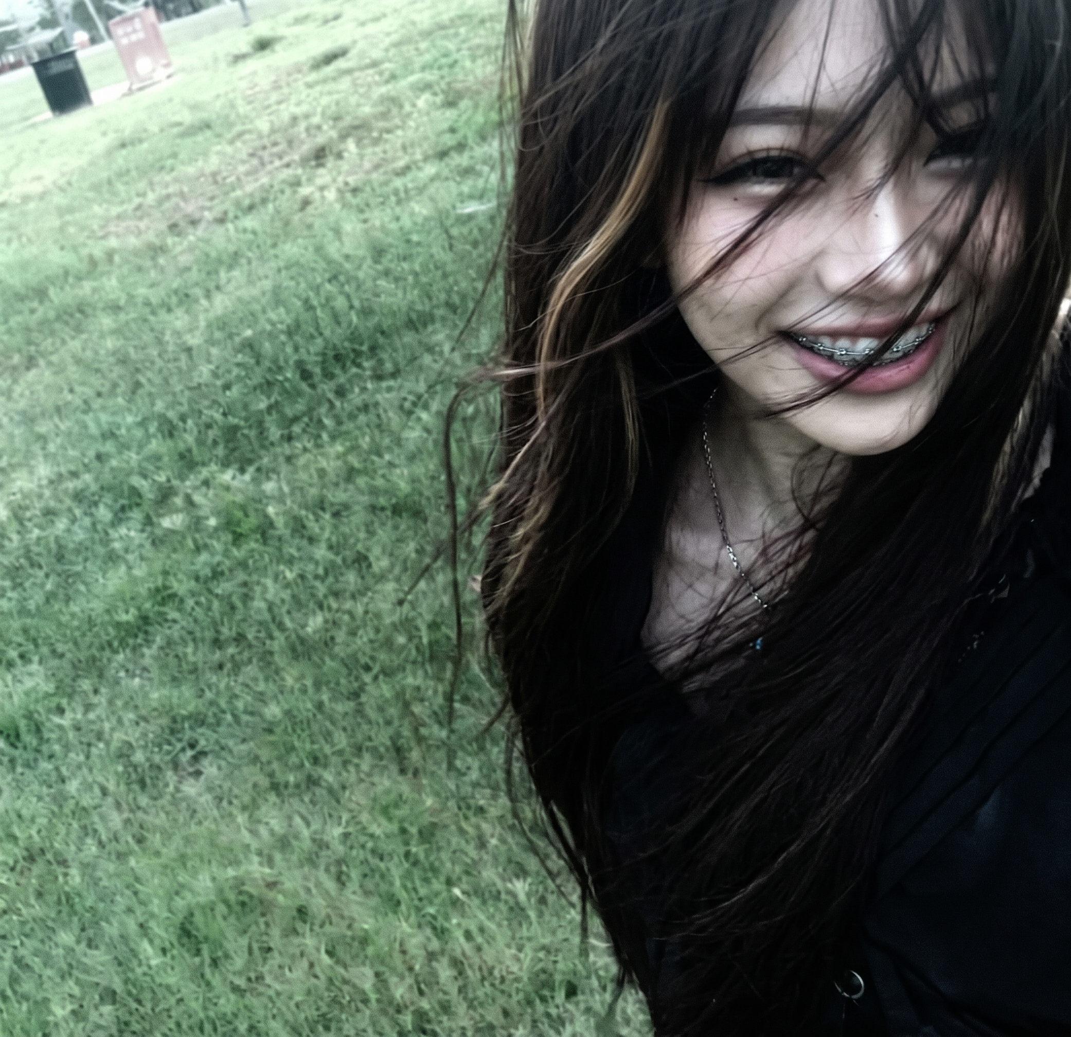 Primela Rose candid outdoor park POV selfie with windswept dark hair, blonde highlights, and signature metallic braces smile in 4K